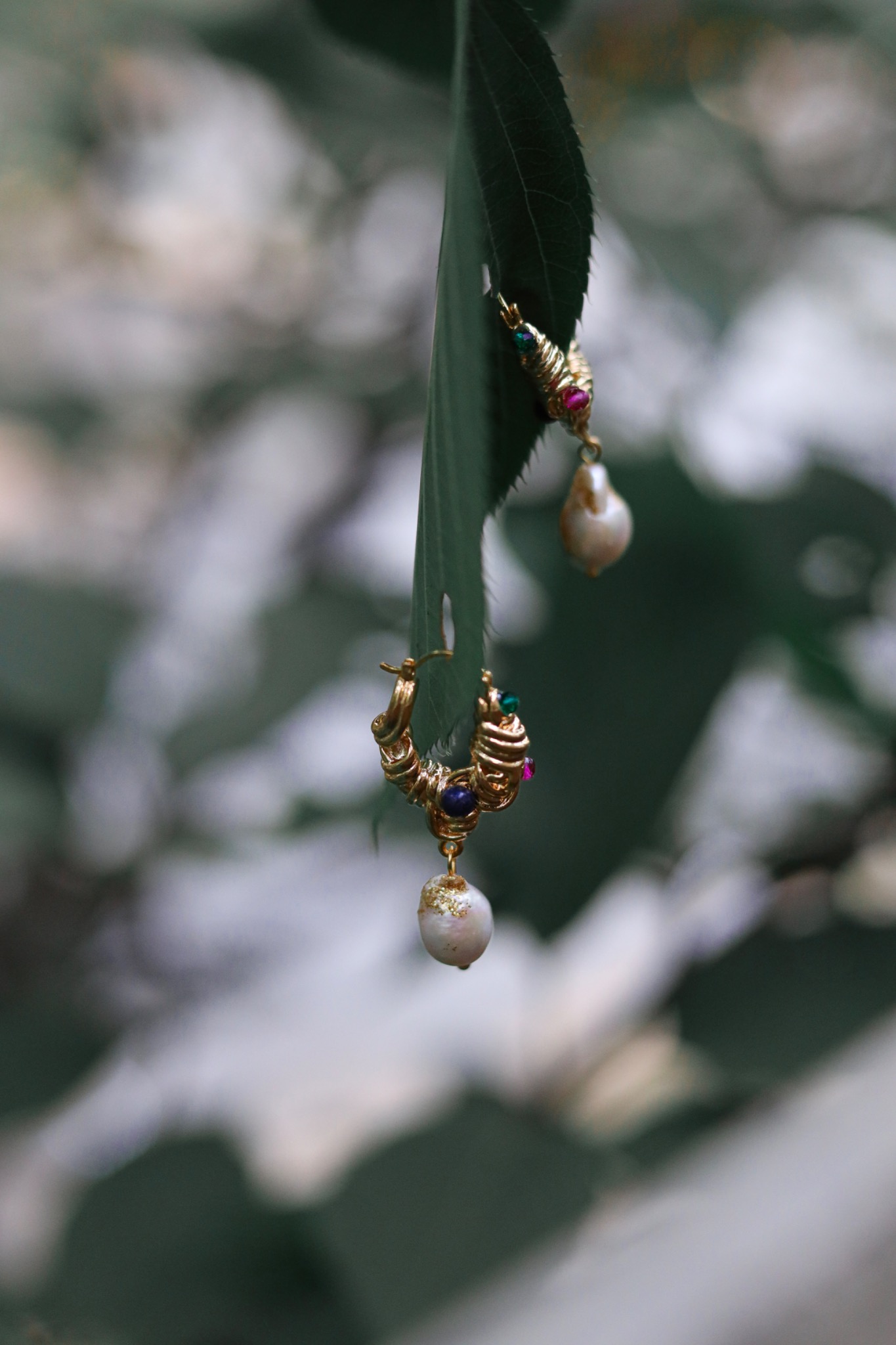 Gold-tone earrings with colorful stones and pearl drops hanging from green leaves